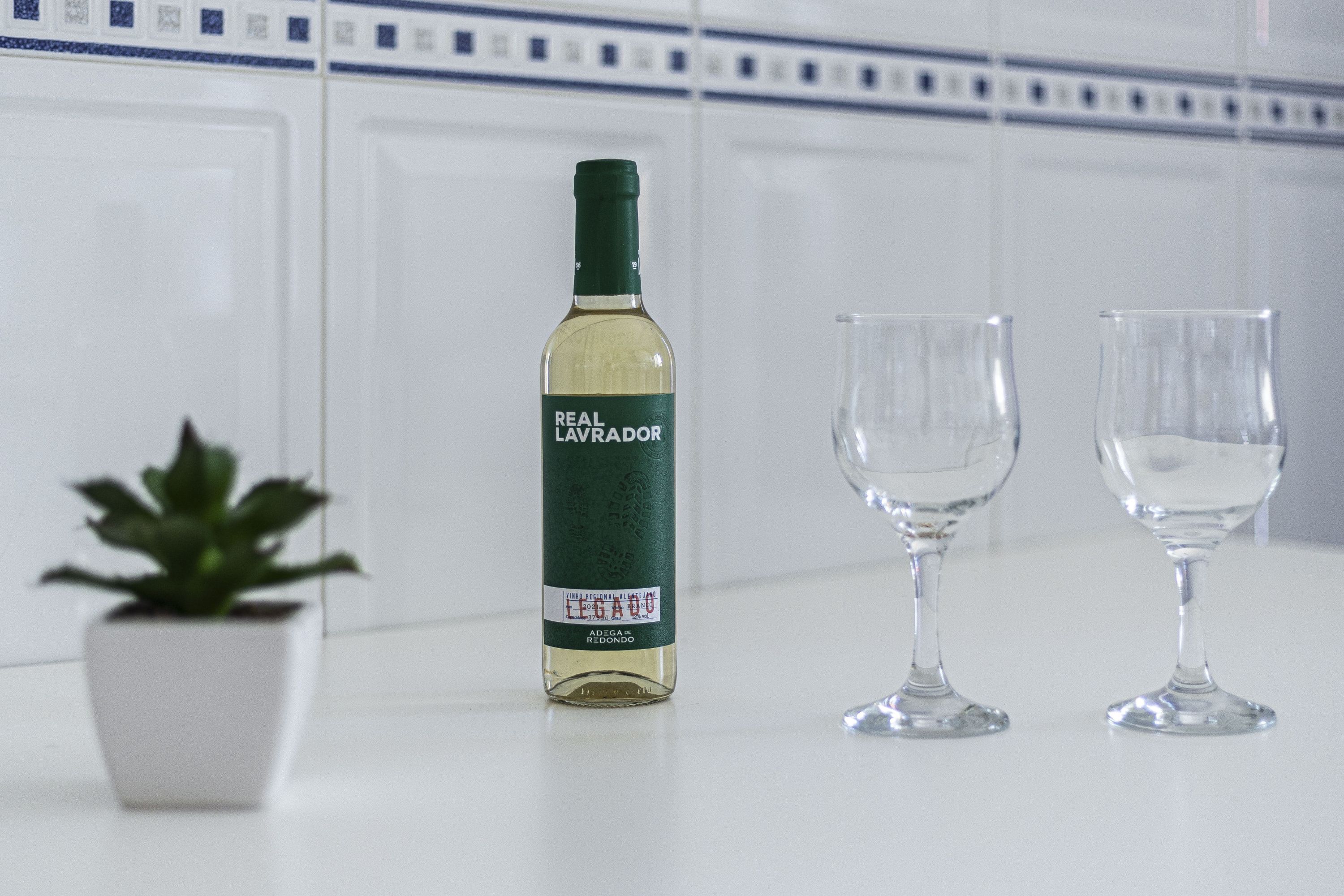 Une bouteille de vin Real Lavador, deux verres à vin vides et une plante en pot, le tout posé sur une surface blanche.