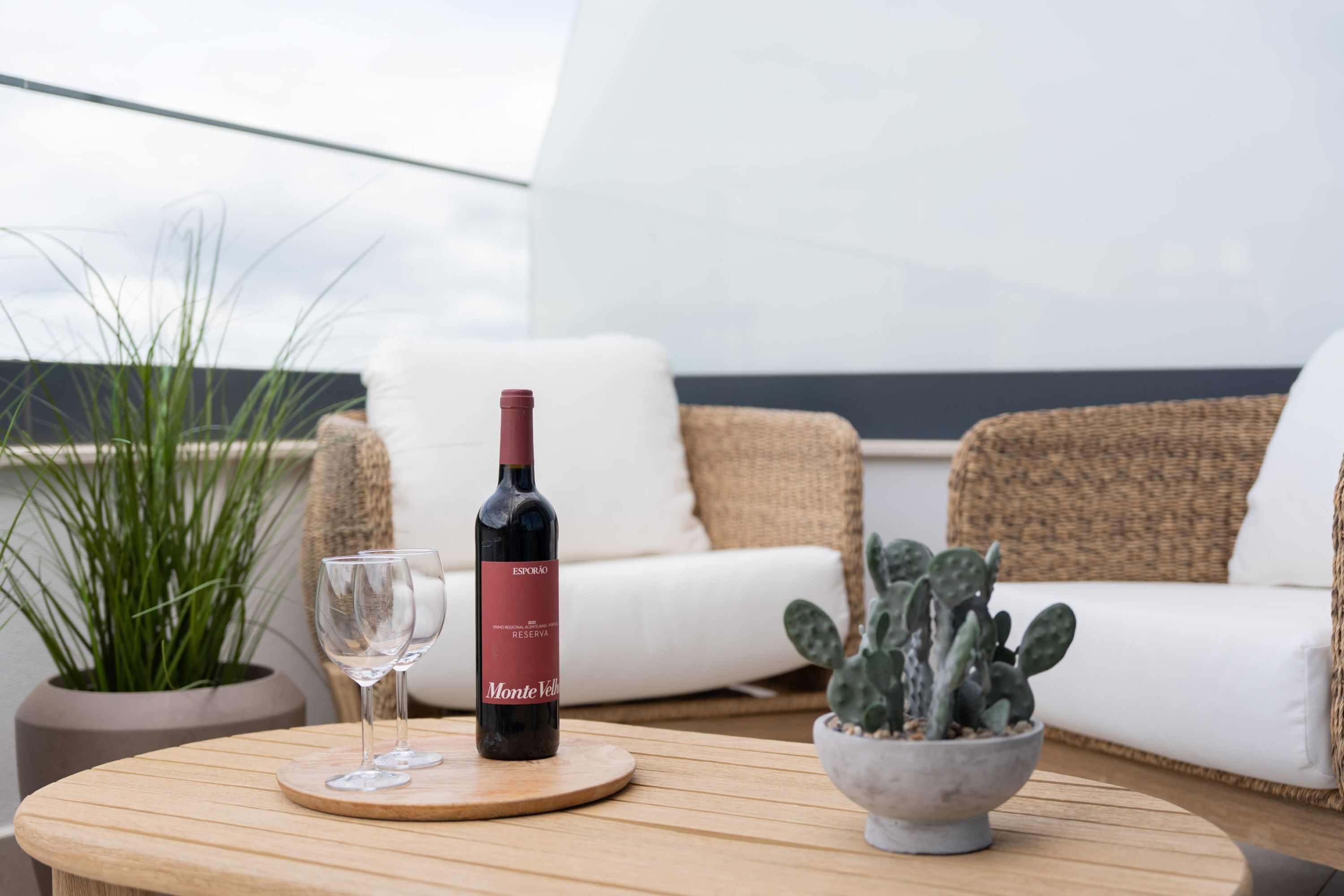 Une table ronde en bois, avec une bouteille de vin, deux verres, une plante en pot et, au fond, un canapé en osier.
