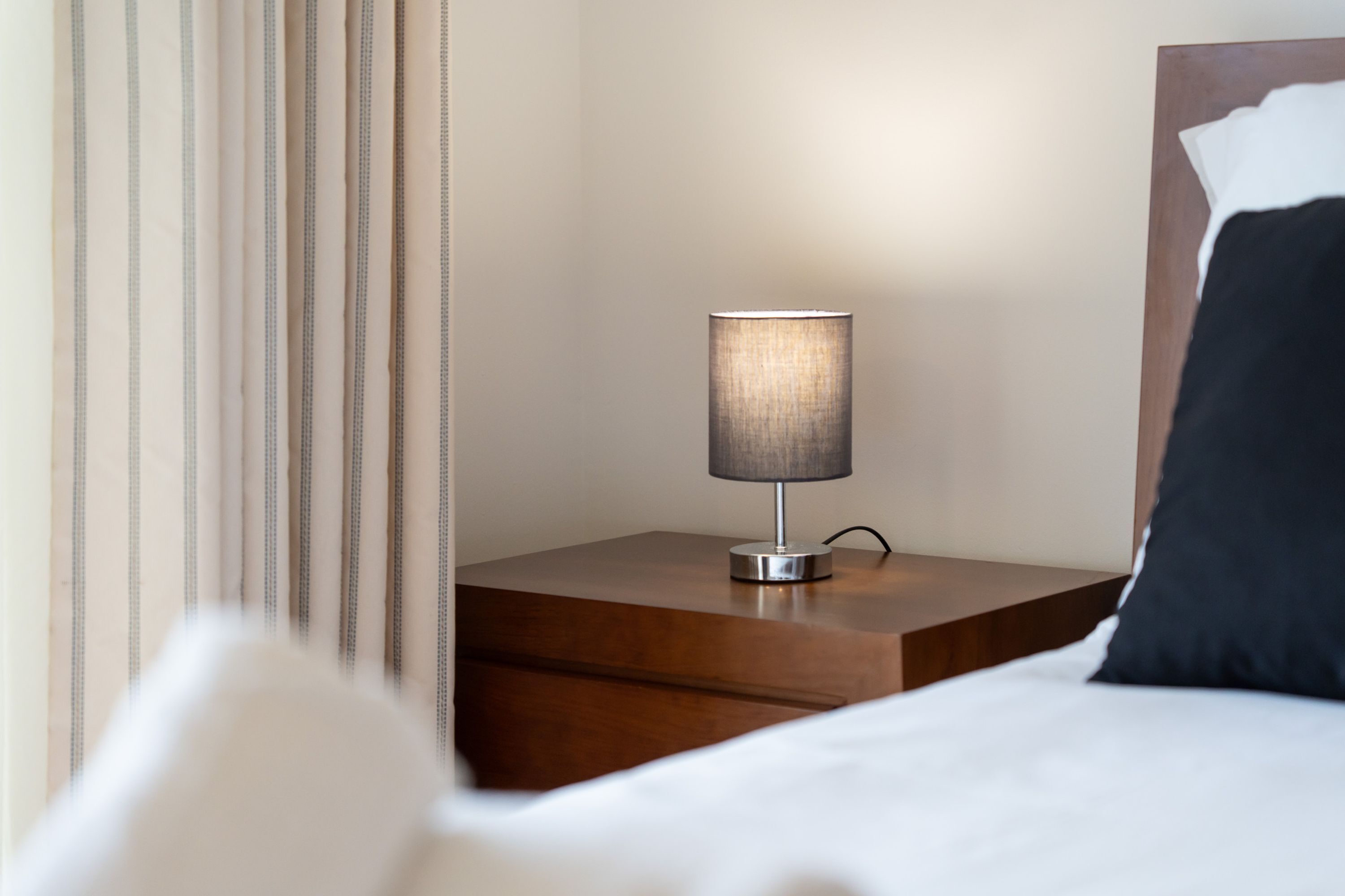 Un lit bien fait, avec une couette blanche et un oreiller noir, une table de chevet en bois avec une lampe, et un rideau à rayures.