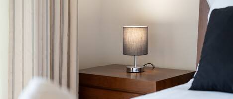 Un lit bien fait, avec une couette blanche et un oreiller noir, une table de chevet en bois avec une lampe, et un rideau à rayures.