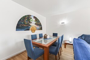Een eetgedeelte met een tafel met glazen blad, blauwe bekleden stoelen, een schilderij van een landschap en een wandlamp.