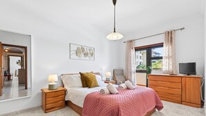 Ein Schlafzimmer mit einem Bett, Nachttischen, einem Fernseher, einer Kommode und einem Fenster mit Vorhängen.