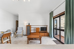 Ein modernes Wohnzimmer mit Schreibtisch aus Holz, einem Fernseher, einem Essbereich mit Tisch und Stühlen sowie einem großen Fenster mit Vorhängen.