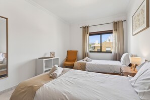 Ein Hotelzimmer mit zwei Betten, einem Sessel, einem Nachttisch mit Lampe, einem Fenster mit Vorhängen und einem gerahmten Bild an der Wand.
