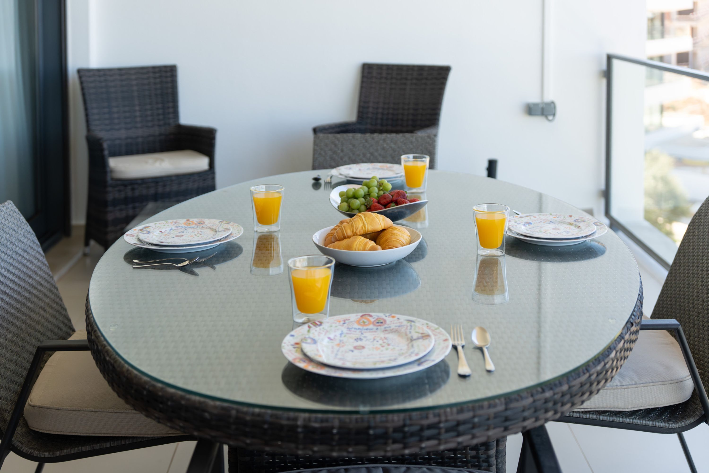 Een ronde glazen tafel met borden, bestek, een schaal croissants en glazen sinaasappelsap.