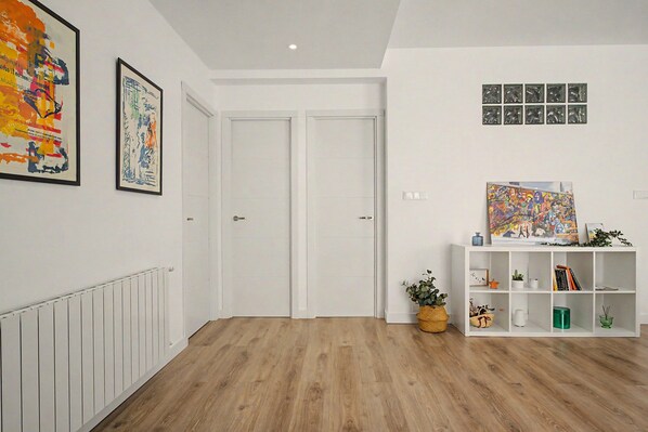 A hallway with a white radiator, two white doors, a white shelving unit with books and decorative items, and a framed abstract painting on the wall.