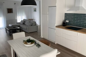 A modern kitchen with white cabinets and a green backsplash, a dining area with a white table and chairs, and a living room with a gray sofa and a black pendant light.