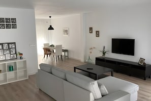 Ein modernes Wohnzimmer mit einer Couch, einem Fernseher, einem Essbereich mit Stühlen und einer weißen Wand mit Kunstwerken.