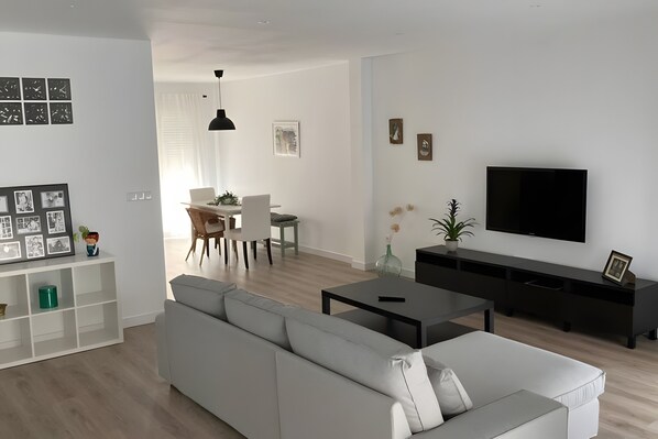 A modern living room with a sofa, a TV, a dining area with chairs, and a white wall with artwork.