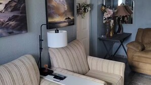 Ett vardagsrum med randiga soffor, ett vitt sidobord, en målning av ett landskap, en lampa, en spegel och en brun fåtölj.