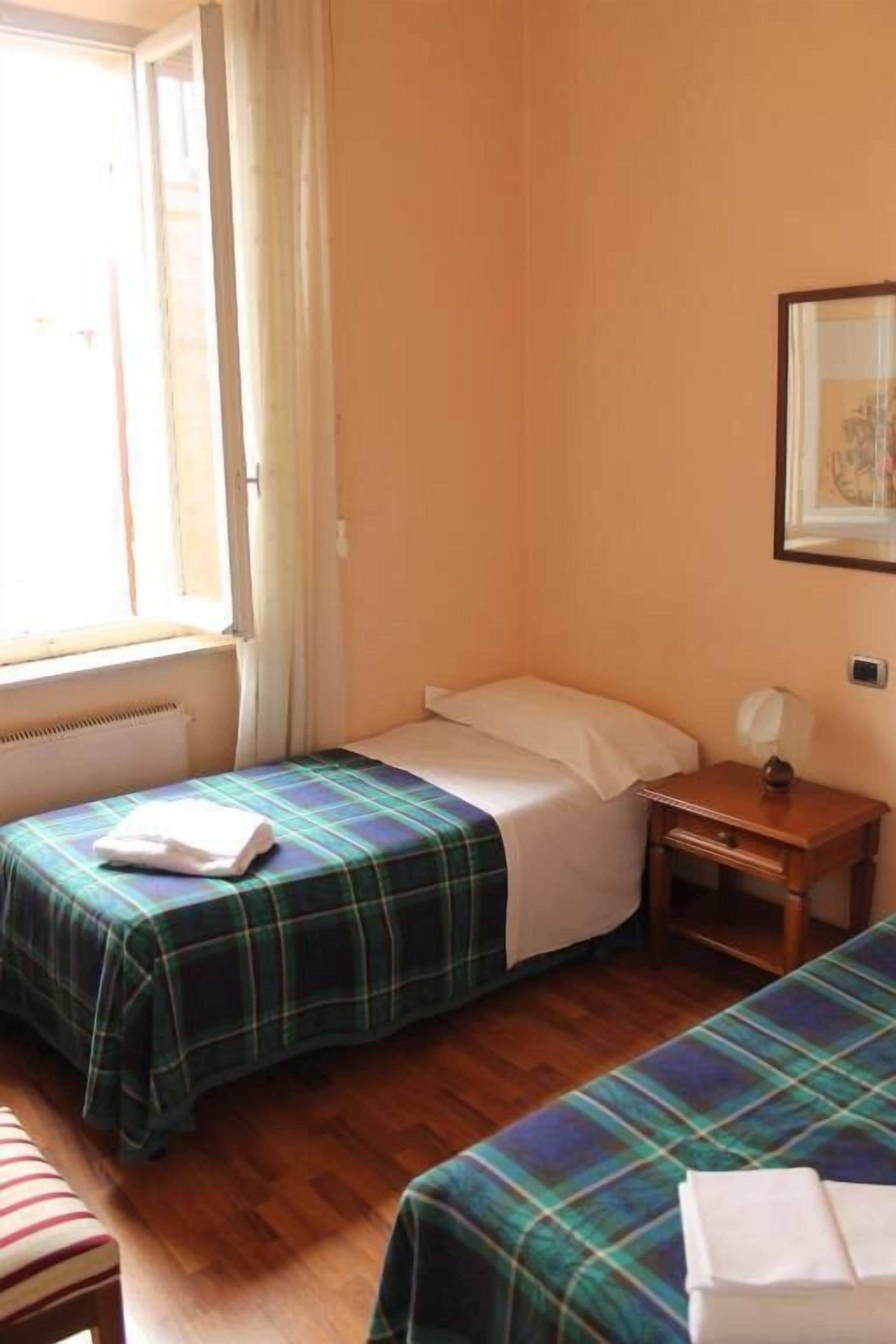A room with two beds, a wooden floor, a window with curtains, a framed picture on the wall, and a small bedside table with a lamp.