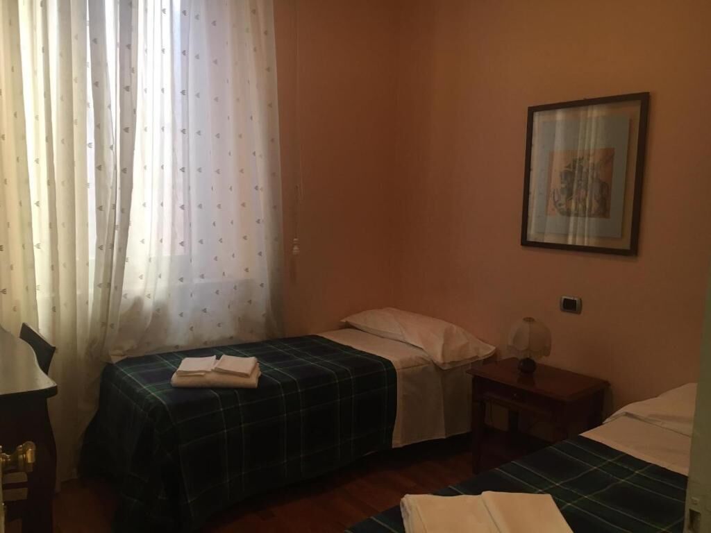 Una habitación con dos camas, un escritorio, una silla, un cuadro en la pared y una ventana con cortinas.