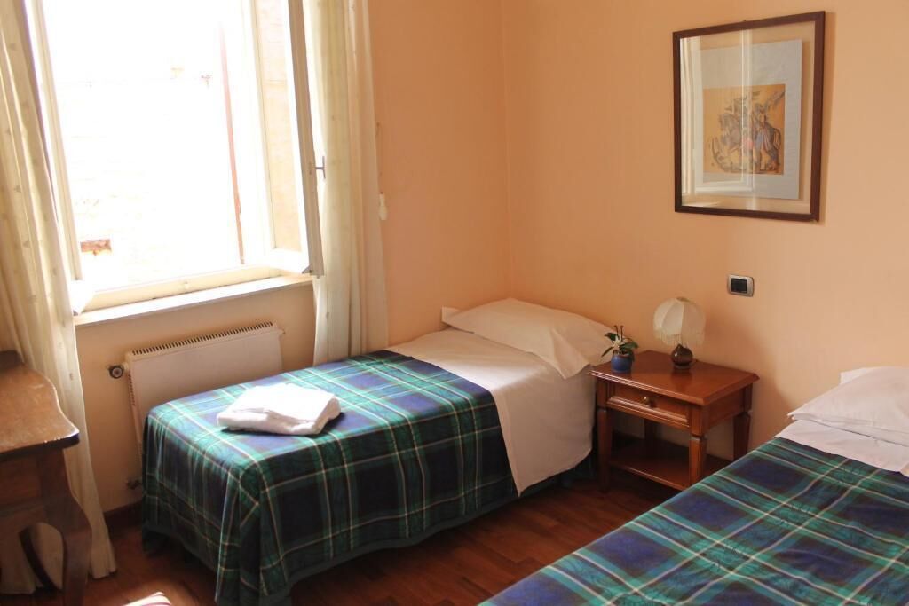 A room with two beds, a window with curtains, a radiator, a small table with a lamp, and a framed picture on the wall.