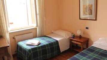 A room with two beds, a window with curtains, a radiator, a small table with a lamp, and a framed picture on the wall.