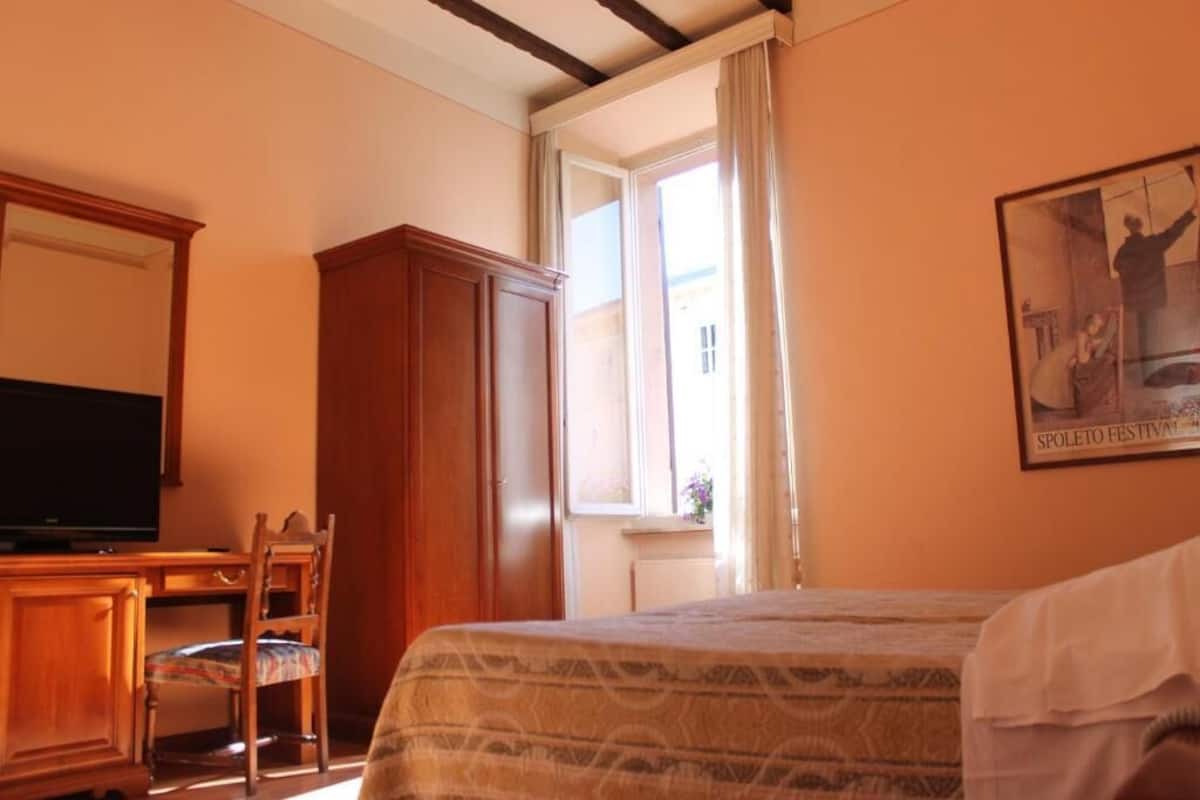 Une chambre d’hôtel comprenant un lit, une télévision, une armoire en bois, un bureau avec une chaise et une fenêtre avec des rideaux.