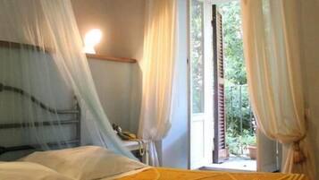 Ein Schlafzimmer mit Bett, einem Fenster mit Vorhängen, einer Nachttischlampe und Blick ins Grüne.