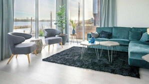 A modern living room with large windows, a teal sofa, two armchairs, a coffee table, and a potted plant. - Pavilion Waterfront 8 (Linden)