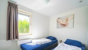 A room with two beds, a window with curtains, a radiator, and a painting on the wall. - Pavilion Waterfront 8 (Linden)