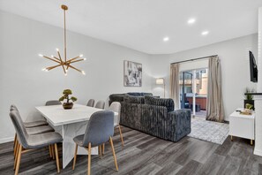 Un salon moderne avec une table à manger blanche, un canapé gris recouvert de tissu et une grande fenêtre avec des rideaux.
