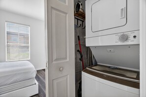 A laundry room with a washing machine, a dryer, and a door leading to a bedroom with a bed and a window with blinds. - Family Retreat: 4BR Home with Private Spa (Kissimmee)