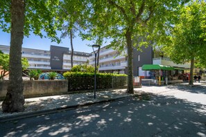 A multi-story building with balconies, a covered outdoor seating area, and a street with trees. - GuestHost - La Casa al Mare APT 406 with Parking (Lignano Sabbiadoro)