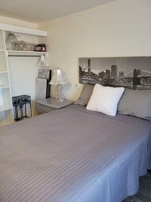 A hotel room with a bed, a nightstand, a lamp, a cityscape painting, and a white shelf with decorative items. - Happy Place Plus (Santa Ana)