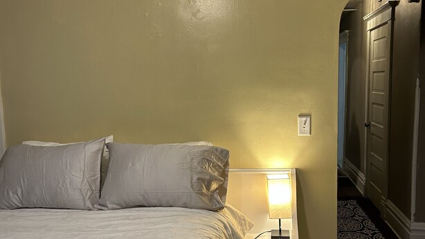 A hotel room with a bed, a nightstand with a lamp, a wardrobe, and a patterned carpet. - Bright apartment in quaint Fargo with refreshing AC (Fargo)