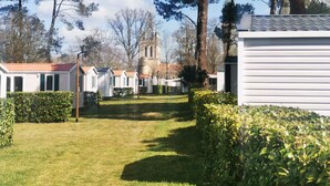 A row of white mobile homes with a grassy yard and a church tower in the background. - 6-person mobile home at a 4-star campground (Lit-et-Mixe)