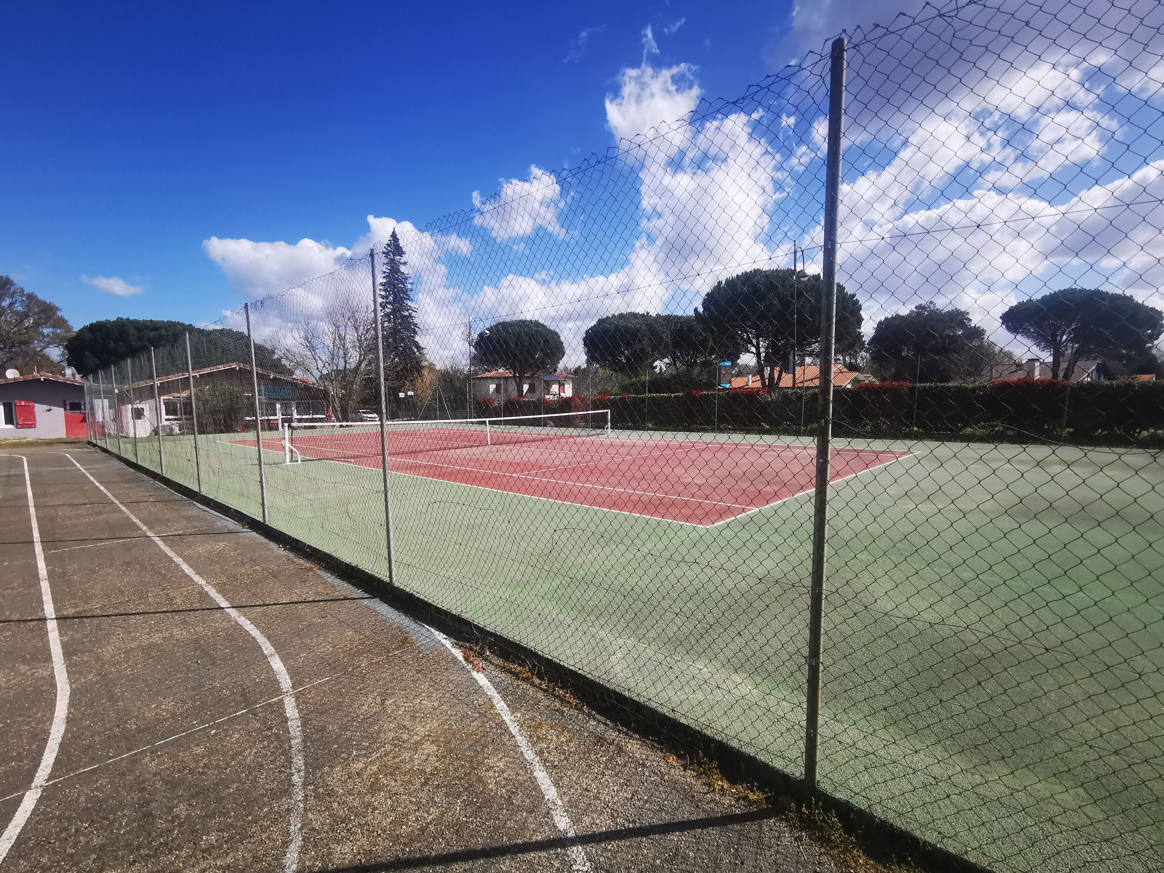 Un court de tennis entouré d’une clôture en treillis métallique, avec un ciel dégagé et des arbres en arrière-plan.