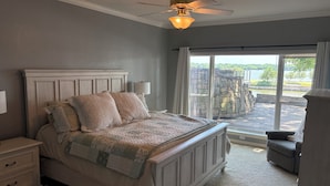 A bedroom with a bed, nightstands, a lamp, a ceiling fan, and a view of a stone wall and water outside. - Waterfront Granbury Getaway — 5BR, Dock & Putting Course (Granbury)