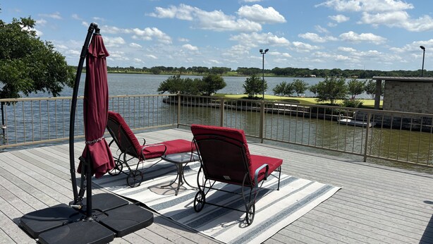 A deck with outdoor furniture, a view of a lake, and a stone building in the background. - Waterfront Granbury Getaway — 5BR, Dock & Putting Course (Granbury)