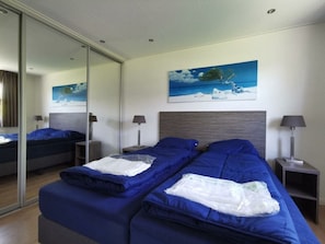 Ein Hotelzimmer mit einem großen Bett, einem kleineren Bett, einem Landschaftsbild und einem Spiegel.