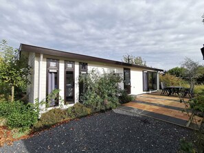 Ein Bungalow mit einer Terrasse, einem kleinen Garten und einer Auffahrt mit Kies.