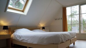 Une chambre avec un plafond en pente, un lit, deux lampes de chevet, une fenêtre avec des rideaux et une vue sur les arbres.