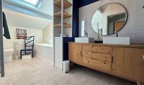 Ein modernes Badezimmer mit Oberlicht, einem Holzbadezimmermöbel, einer weißen Badewanne und einem blauen Sessel.