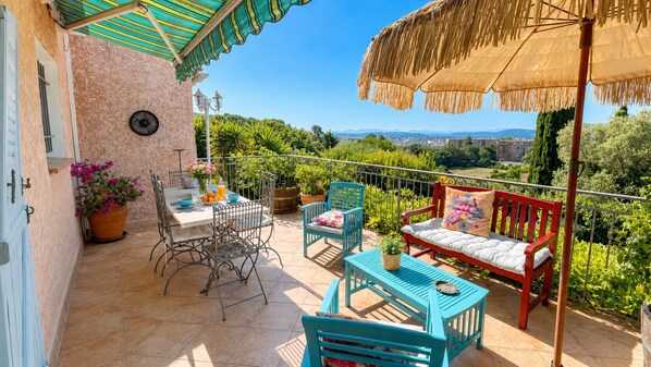 Ein Balkon mit Tisch, Stühlen und einer Sitzecke, umgeben von Blumentöpfen und Blick auf die Landschaft.
