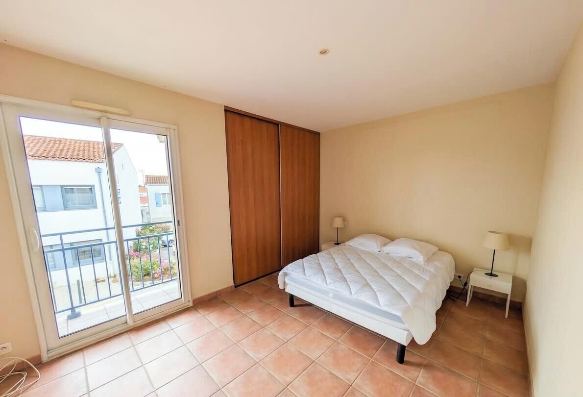 Ein Schlafzimmer mit Bett, Nachttisch, Lampe, einer Schiebetür zum Balkon und Blick auf Gebäude.