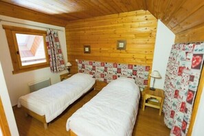 Una habitación tipo cabaña con dos camas individuales, una ventana con cortinas, techo de madera y una mesita con lámpara.