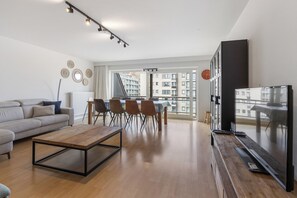 A modern living room with a sofa, coffee table, dining table with chairs, a TV, and a large window with a view of buildings. - 2 bedroom accommodation in Oostduinkerke (Oostduinkerke)