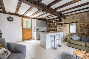 Eine rustikale Küche mit Steinwand, Holzbalken, einer weißen Kochinsel mit Barhocker, einem grauen Sofa und einem grünen Sofa mit Kissen.