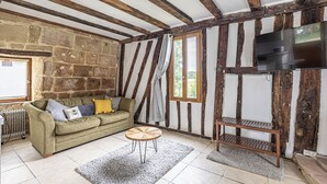 Ein Zimmer mit freiliegenden Holzbalken, einer Steinwand, einer Couch mit Kissen, einem runden Couchtisch, einem Fernseher und einem Fenster mit Vorhängen.