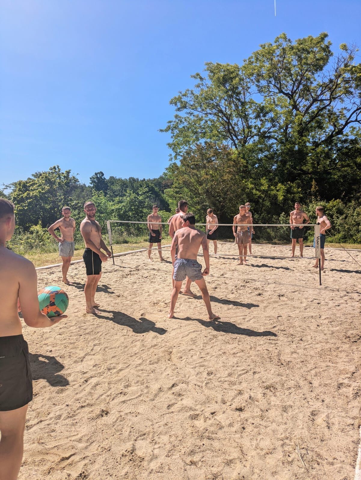 A group of people playing beach volleyball on a sandy court with trees in the background.