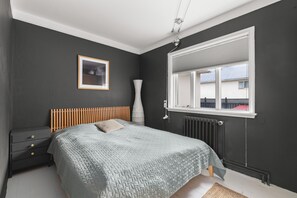 Ein Schlafzimmer mit Bett, Nachttisch, Fenster, Heizkörper und einem gerahmten Bild an der Wand.