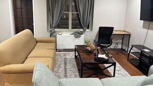 Una sala de estar con un sofá beige, un sofá gris, una mesa de centro de madera con una guitarra, una silla de oficina negra, un radiador blanco y un televisor montado en la pared.