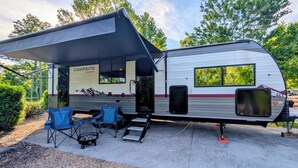 A camper with a covered patio area, two blue folding chairs, a grill, and a sign reading 'Campsite Reserve.' - Cozy riverside camper in peaceful Townsend with AC (Townsend)