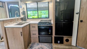 A compact kitchen with a built-in oven, microwave, and refrigerator, a window with a view of greenery, and a small sink. - Cozy riverside camper in peaceful Townsend with AC (Townsend)