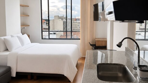 Quarto de hotel moderno com uma cama, área de cozinha com pia, televisão e vista para a cidade.
