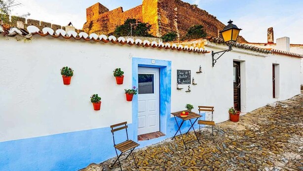 Un bâtiment blanc avec une porte bleue, des plantes en pot, une table avec des chaises et une rue pavée.