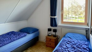 Ein Schlafsaalzimmer mit zwei Betten, einem Fenster mit Blick auf Bäume, einem Heizkörper und einem kleinen Nachttisch aus Holz mit zwei Lampen.