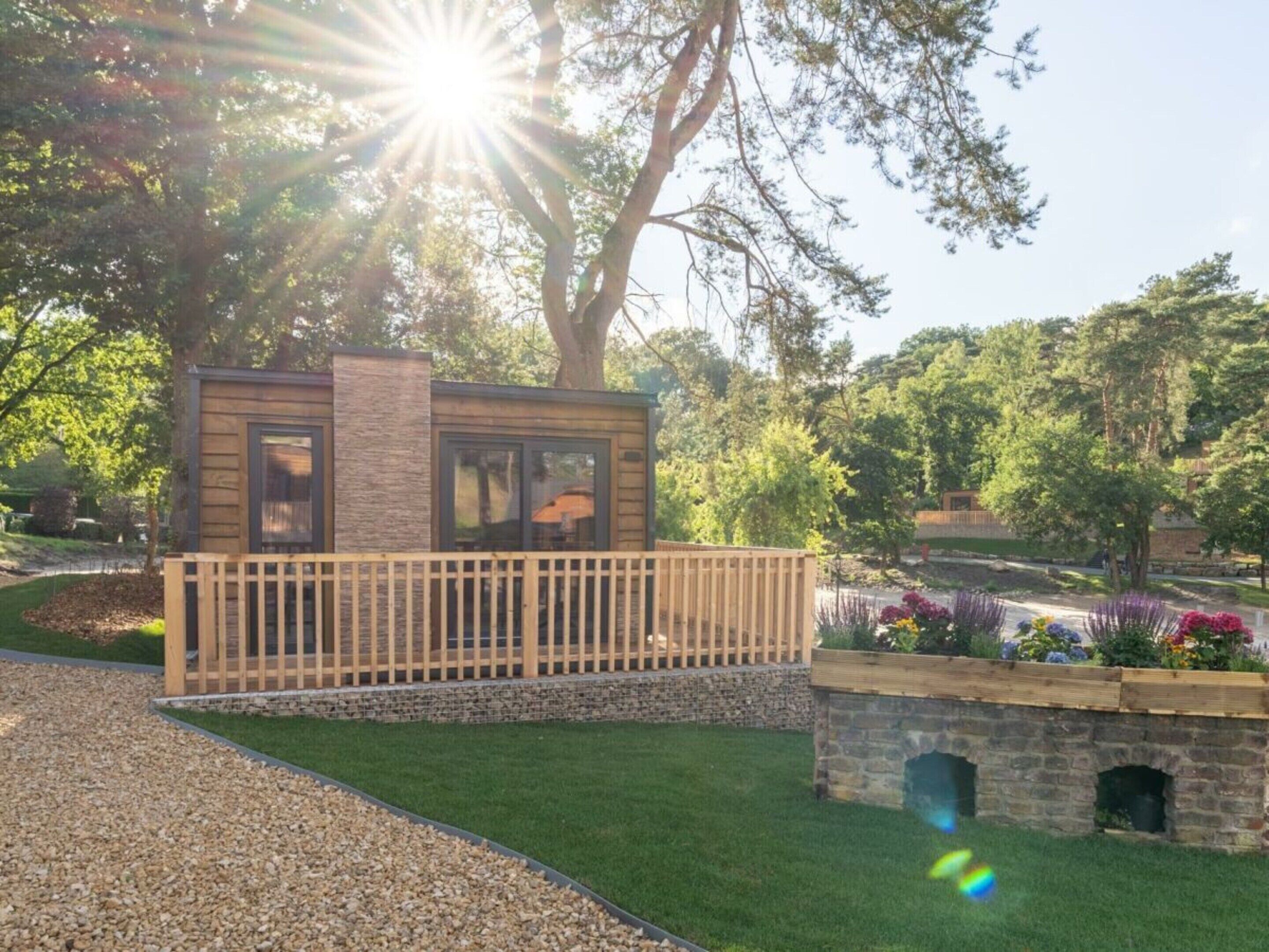 Ein kleines, modernes Gebäude im Blockhausstil mit Holzterrasse und Steinwänden, umgeben von einem gepflegten Garten und Bäumen.